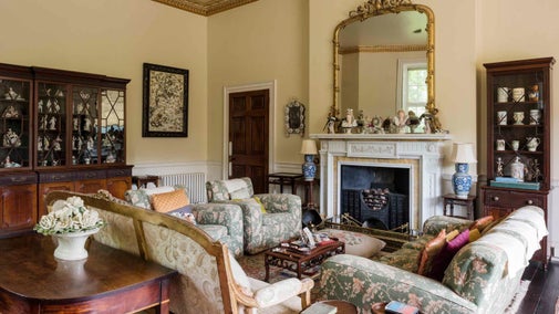 The Drawing Room at Greenway in Devon. Floral patterned sofas and chairs face each other surrounding a marble fireplace with large gold mirror above. Cabinets filled with ceramics line the walls.